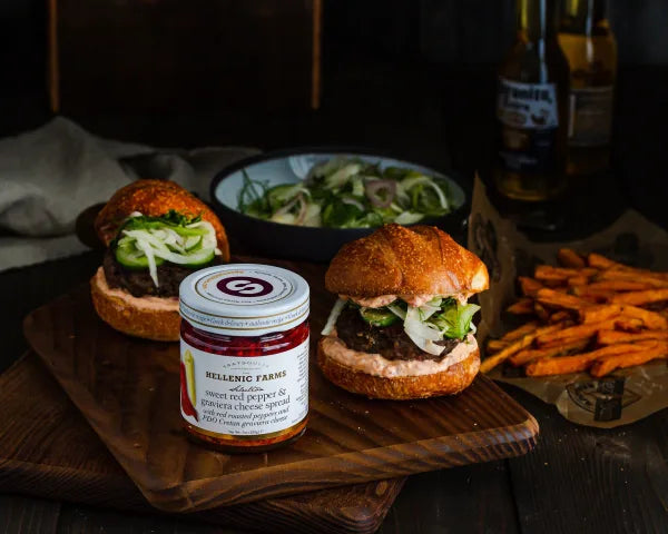 Two burgers with a jar of Hellenic Farms spread on a wooden board with fries and salad in the background.