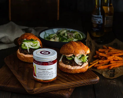 Two burgers with a jar of Hellenic Farms spread on a wooden board with fries and salad in the background.