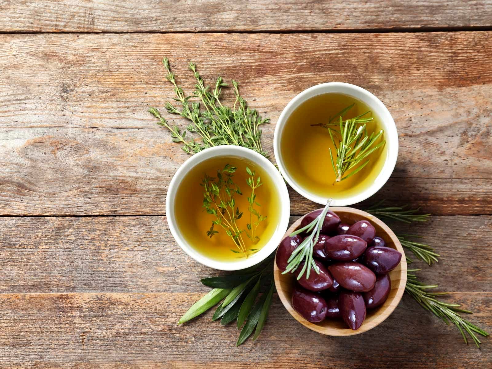 Greek extra virgin olive oil with thyme and rosemary beside a bowl of Kalamata olives on a rustic wooden table